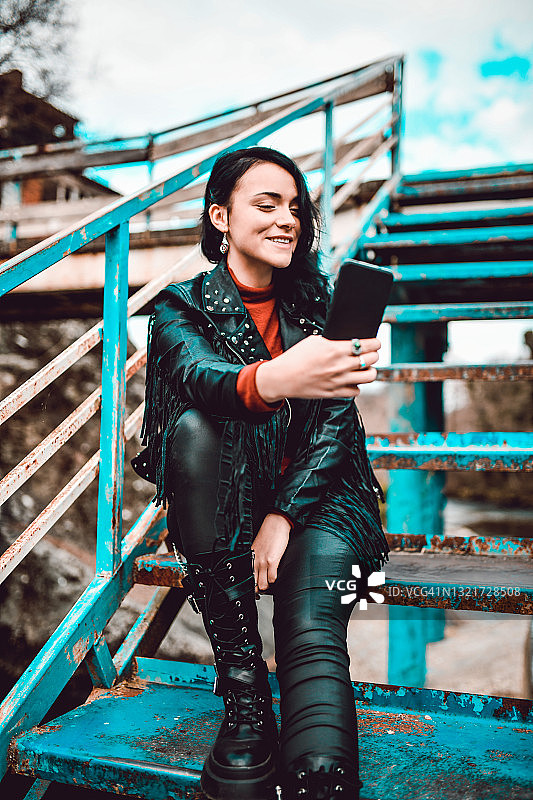 一位微笑的哥特式女性在锈桥上用智能手机自拍图片素材