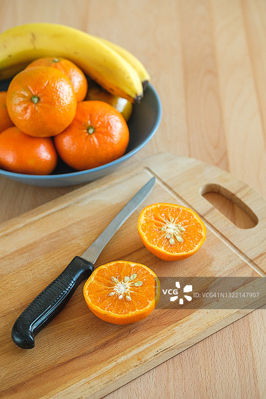 把带骨的橘子切成两半，放在砧板上。旁边是一盘水果、橘子和香蕉，背景是一张木制餐桌。素食、纯素和生食。有机农家产品的概念。图片素材