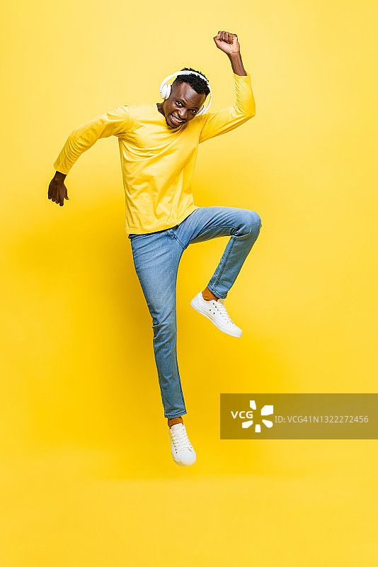 快乐的年轻非洲人戴着耳机听音乐和跳跃与手在黄色孤立的演播室背景图片素材