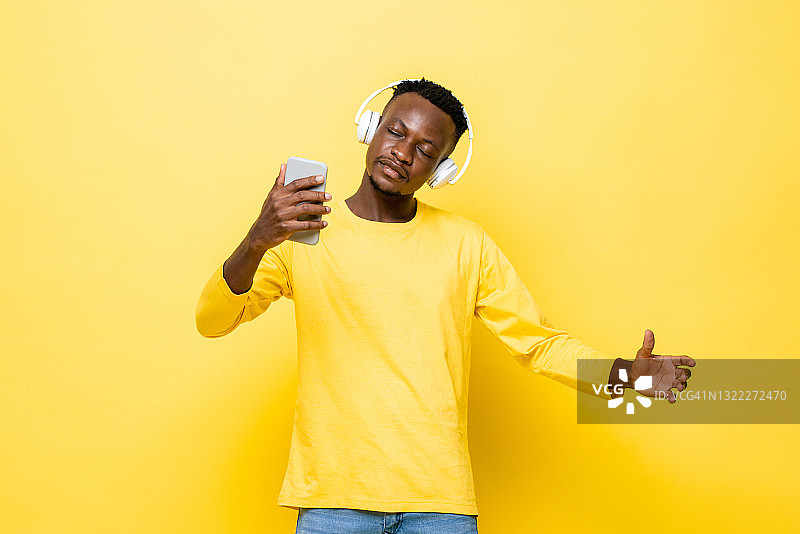 放松年轻的非洲男子戴着耳机听在线音乐从手机在孤立的黄色演播室背景图片素材