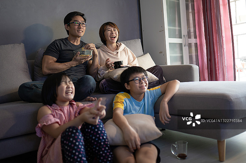 亚裔华人一家人坐在家里的沙发上看电视。图片素材
