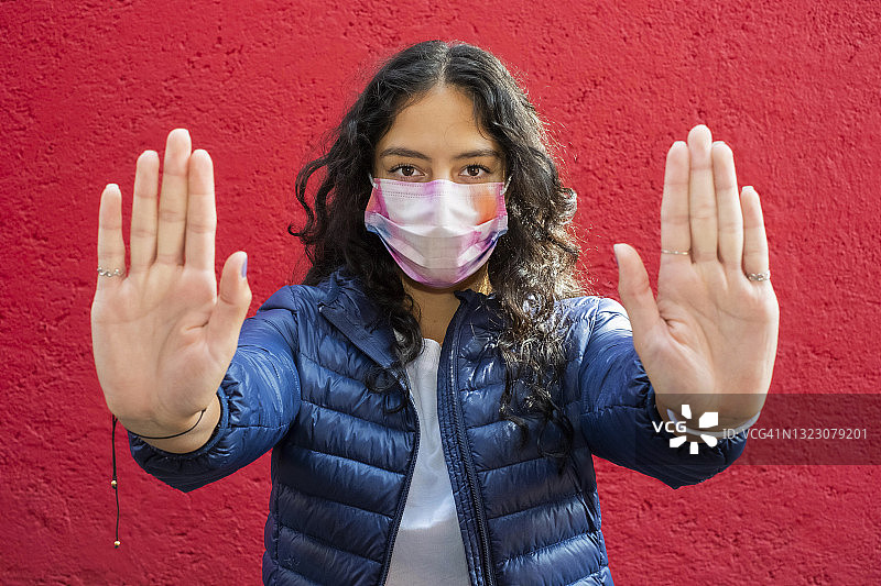 年轻的拉丁妇女戴着口罩，看着摄像机，并手势停止用双手，红色的背景图片素材