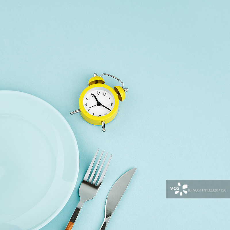 间歇性禁食、节食、午餐时间概念。黄色闹钟，空盘子和餐具在蓝色背景。图片素材