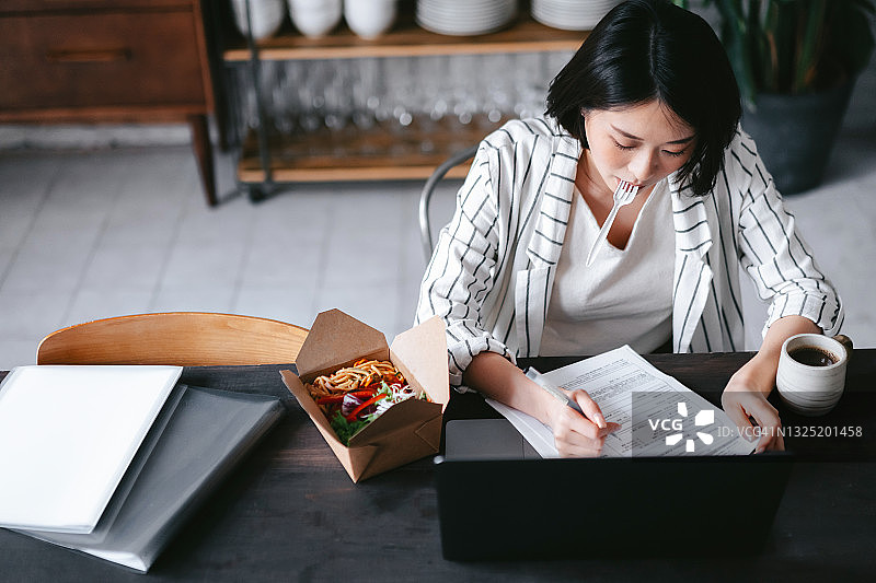 忙碌的年轻亚洲女性一边吃着健康的外卖沙拉盒饭，一边在家用笔记本电脑工作，在家里的客厅里处理文件。远程工作概念图片素材