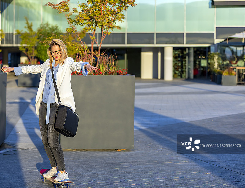 一个年轻漂亮的女人穿着商务套装在城市街道上带着她的滑板冲向办公室。图片素材