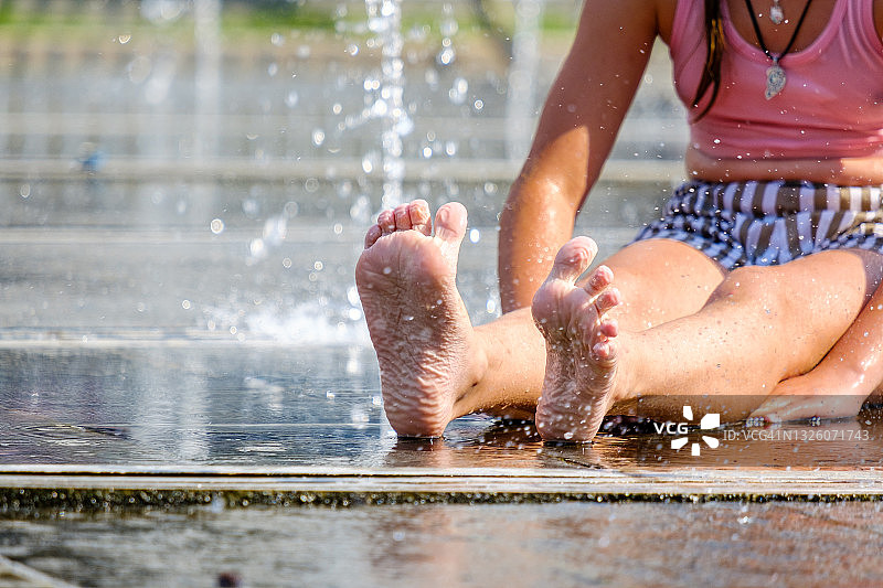 一个小女孩坐在街头喷泉的地板上，被水花溅起。图片素材