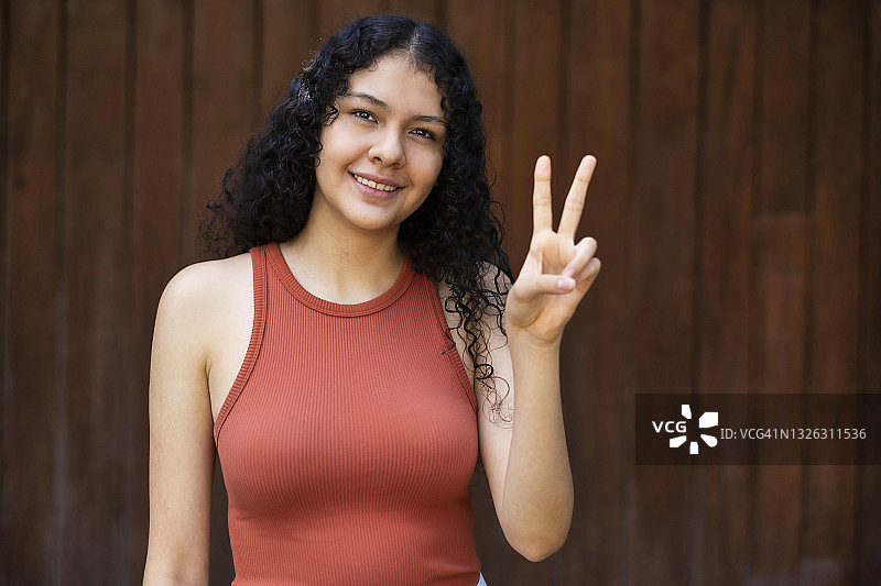 年轻的拉丁美洲妇女与卷曲的头发微笑，看着相机，并给出和平的手势图片素材