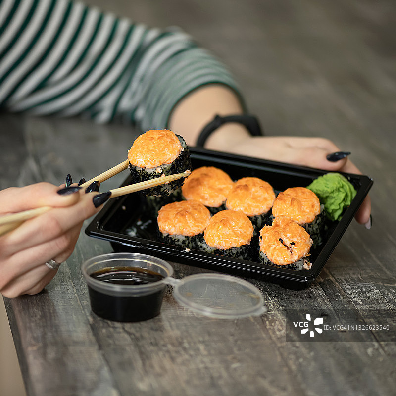 女人用筷子从黑色塑料容器吃热烤寿司卷。外卖日本料理概念。近距离射杀。软焦点图片素材