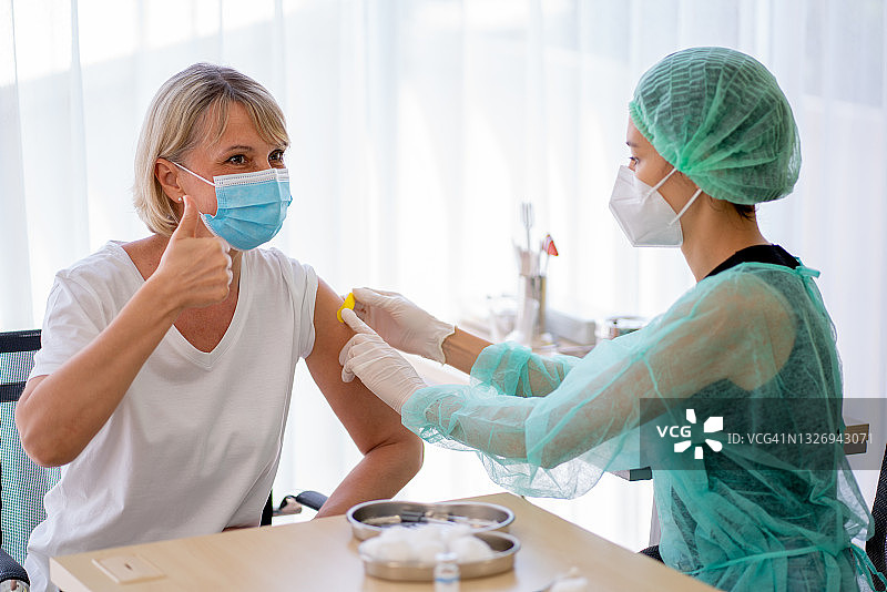 佩戴卫生口罩、身着绿色制服的护士或医务人员为年长女性注射Covid-19疫苗，年长女性竖起大拇指图片素材