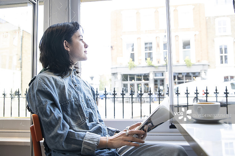 一个年轻的女人坐在咖啡馆里，拿着书透过窗户看图片素材
