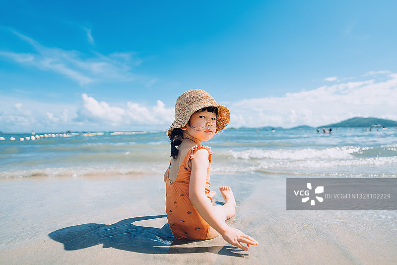 可爱的亚洲小女孩，戴着草帽，坐在海边，被海浪拍打着。在阳光明媚的夏日，在海滩上玩得很开心图片素材