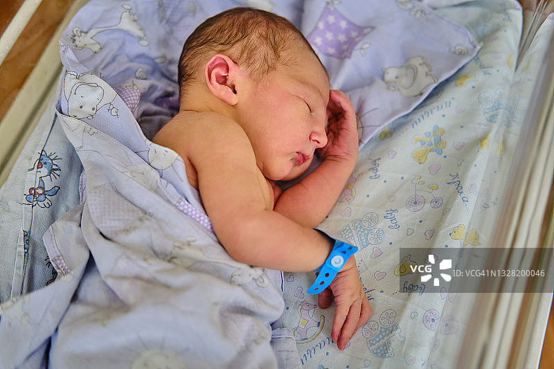 一个胳膊上戴着产科医院手镯的新生儿正在婴儿床里睡觉。一个刚出生的孩子躺在一个透明玻璃后面的病床上图片素材