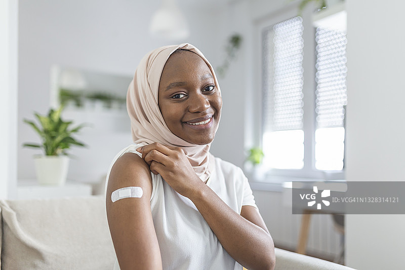 一名非洲穆斯林妇女在接种疫苗后微笑的肖像。妇女按住她的衬衫袖子，并显示她的手臂与绷带接受疫苗接种后。图片素材