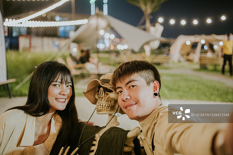 自拍，年轻的泰国夫妇看智能手机为可爱的自拍约会在万圣节概念展在晚上露营咖啡馆库存照片图片素材