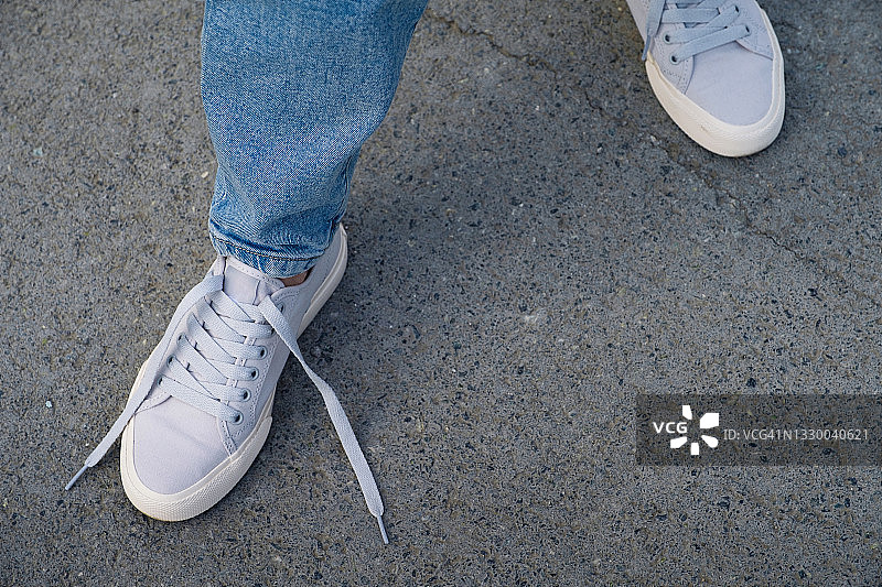 女孩的脚穿着新的干净的现代运动鞋和牛仔裤，一条鞋带是解开的。一个穿着运动鞋的女人走在人行道或柏油路上。时尚时尚的生活方式。文本空间的拷贝。图片素材