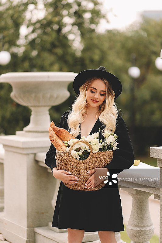 一个漂亮的漂亮可爱的年轻女人，一头金发，带着一顶帽子和一件优雅的黑色连衣裙，手里拿着一个装着鲜花和面包的柳条袋，在一个整洁的公园里散步，在剧院和城市建筑附近的植物和树木之间图片素材