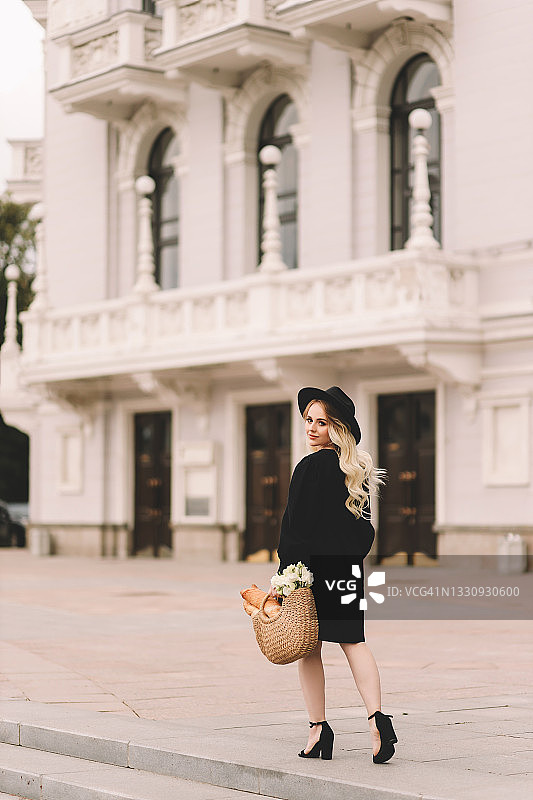 一个漂亮的漂亮可爱的年轻女人，一头金发，带着一顶帽子和一件优雅的黑色连衣裙，手里拿着一个装着鲜花和面包的柳条袋，在一个整洁的公园里散步，在剧院和城市建筑附近的植物和树木之间图片素材