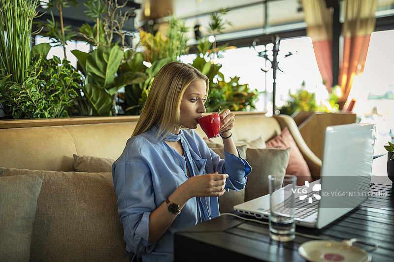 一个年轻美丽的女人正在使用她的笔记本电脑，而坐在现代餐厅和清凉的热饮。图片素材
