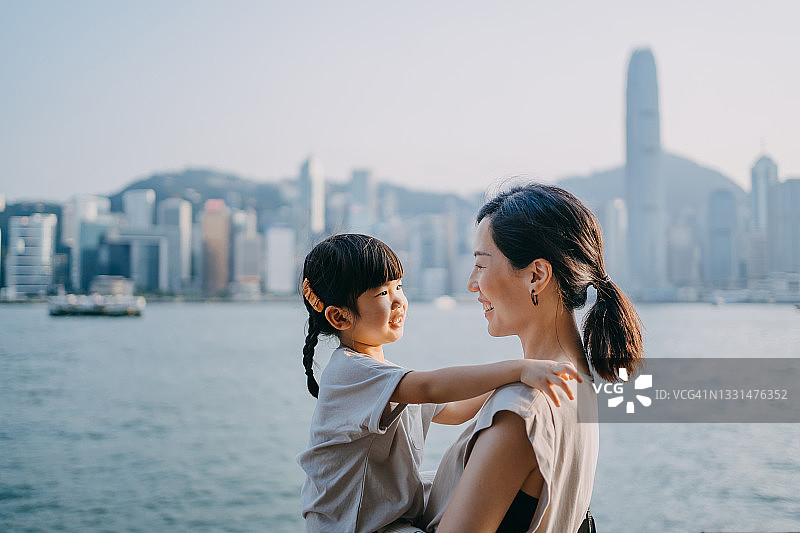 年轻可爱的亚洲妈妈抱着可爱的小女儿，站在维多利亚港的海滨长廊上，谈笑风生。一起分享亲密的时间图片素材