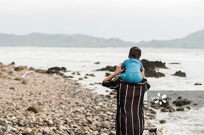 东南亚母亲背着她的儿子，在白天走在海滩上图片素材
