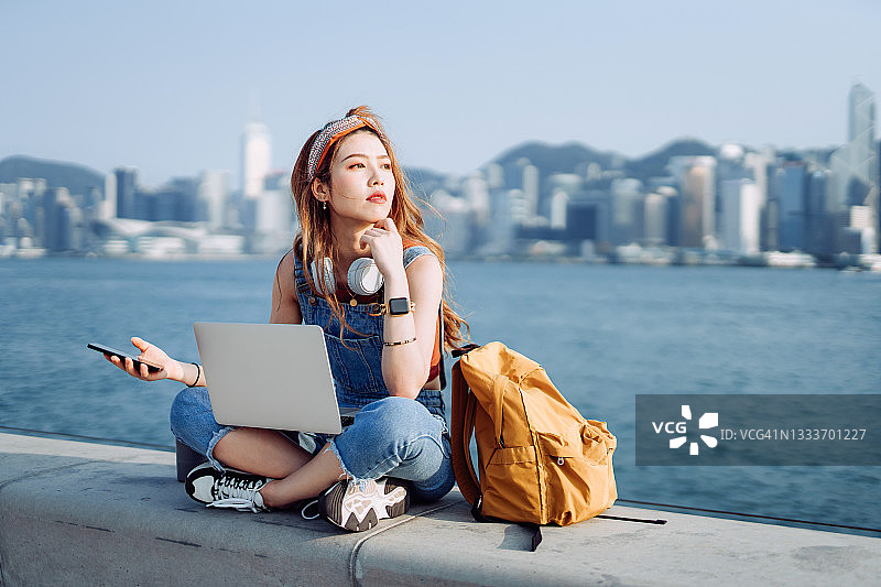 有思想的年轻亚洲女人盘腿坐在人行道上，对着城市的天际线。她脖子上戴着耳机，用智能手机和笔记本电脑工作。青少年的生活方式和科技图片素材