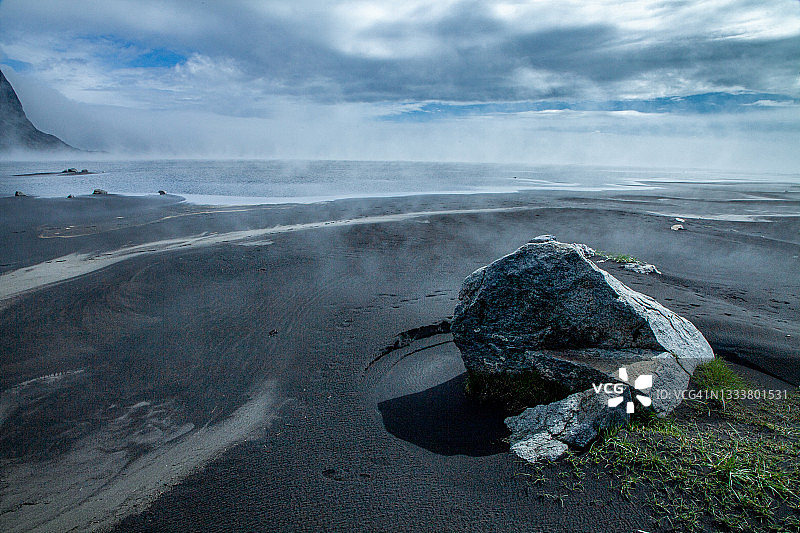 岩石和黑色沙滩的POV与雾升起的海洋和引人注目的山脉和云填充天空的背景。图片素材