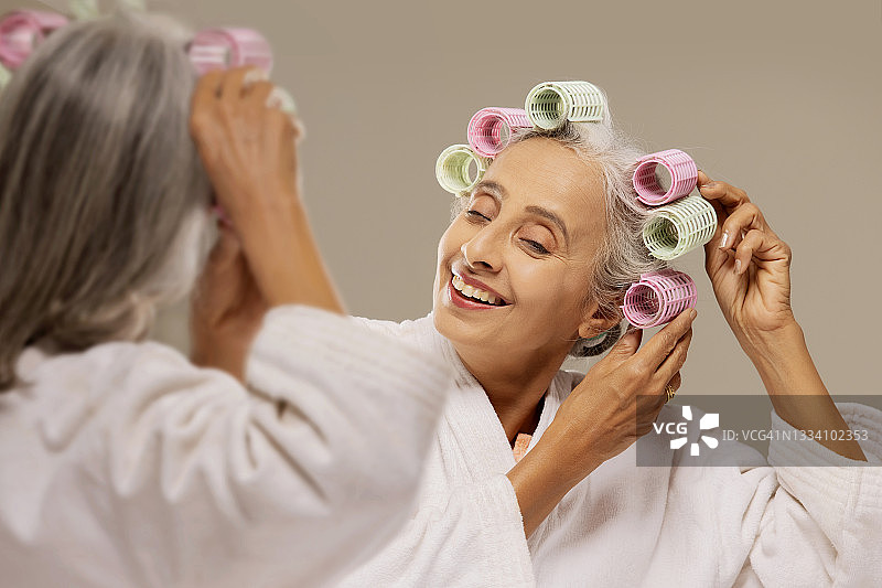 一位老妇人在镜子前用卷发器做发型。图片素材