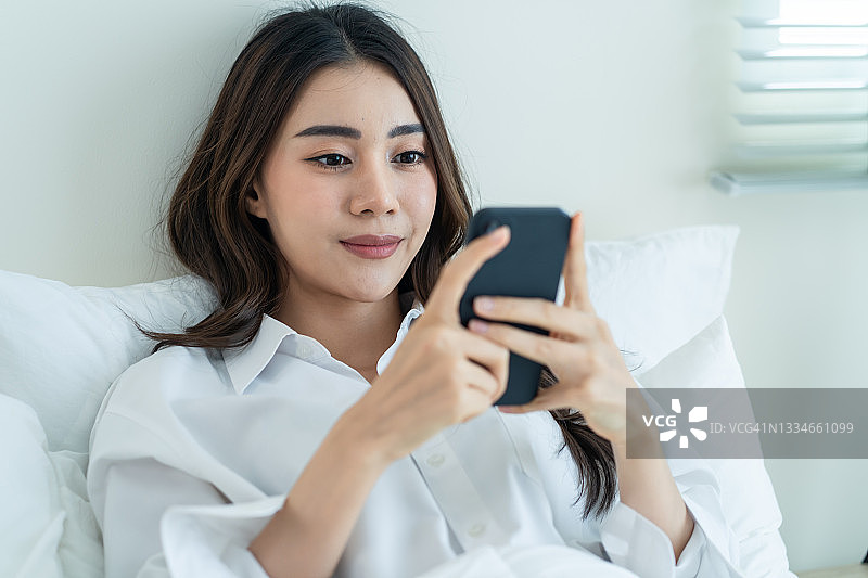 亚洲美女早上在家里的床上用手机聊天。快乐随意的漂亮女性躺在卧室享受假日周末，在家里用智能手机和科技交流。图片素材