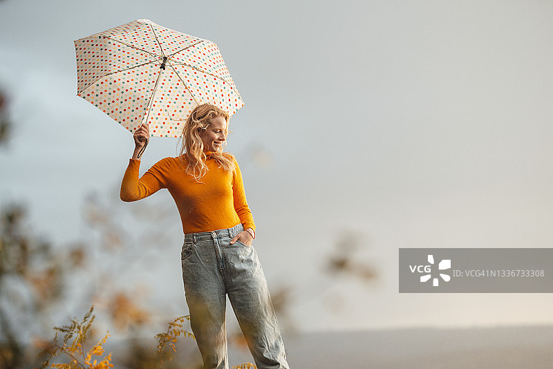 微笑的金发女人手持伞，手插在口袋里图片素材