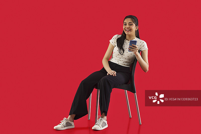 一名年轻女子拿着智能手机坐在红色背景的椅子上。图片素材