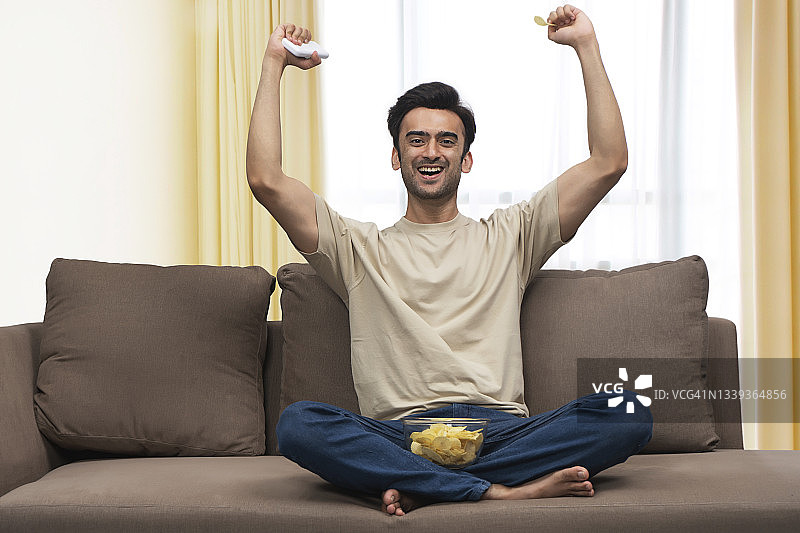 一个年轻人在家里庆祝比赛胜利，腿上放着零食。图片素材