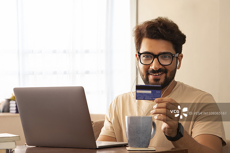 一名男子带着笔记本电脑，戴着耳机，展示着信用卡，面带微笑。图片素材