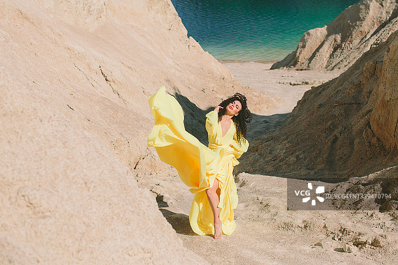 一名年轻女子站在采石场的岸边，靠近沙石和湖泊，身穿黄色夏装，对着镜头摆姿势图片素材