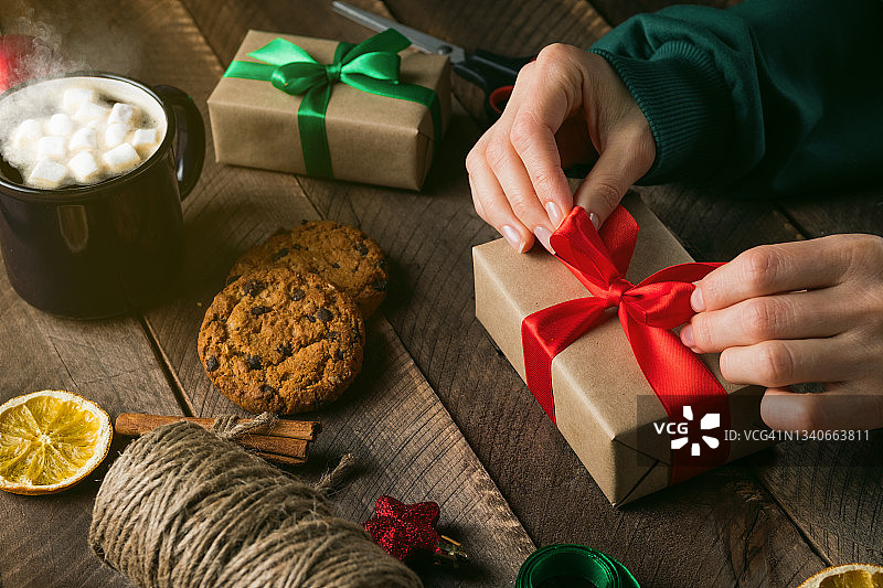 一个穿绿色毛衣的女孩或妇女拿着并系上圣诞礼物。全家准备过节的概念，平安夜快乐。木桌的背景上放着热咖啡和棉花糖，还有巧克力饼干。图片素材