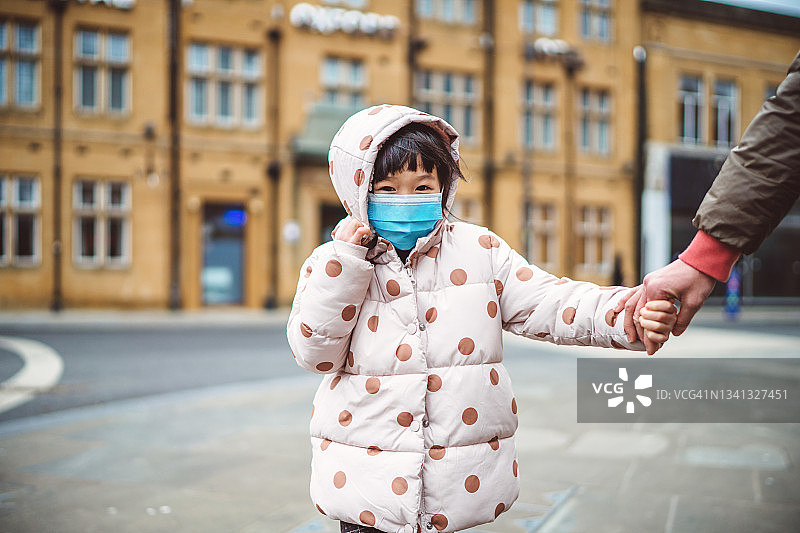 可爱的小女孩戴着防护面具，对着镜头快乐地笑着，和她的妈妈在街上散步图片素材