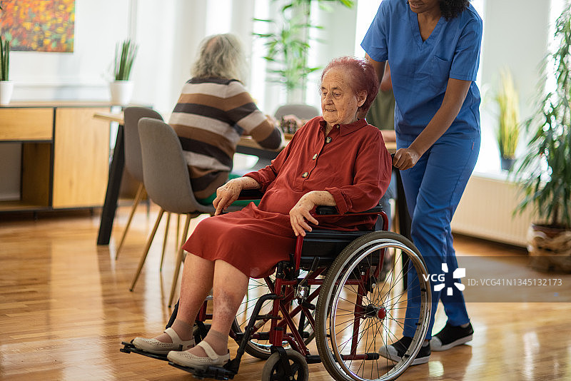 坐在轮椅上的老年妇女在女护士的帮助下穿过养老院图片素材