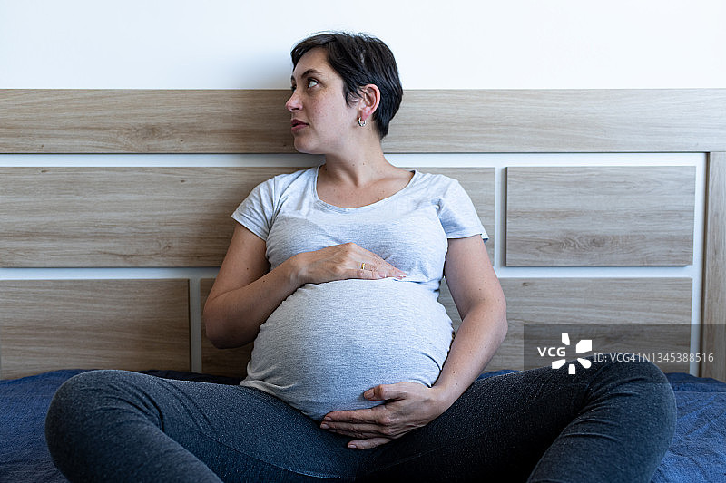 非常怀孕的短发女人坐在她的床上，看着她的右边，双手抓着她的肚子。图片素材