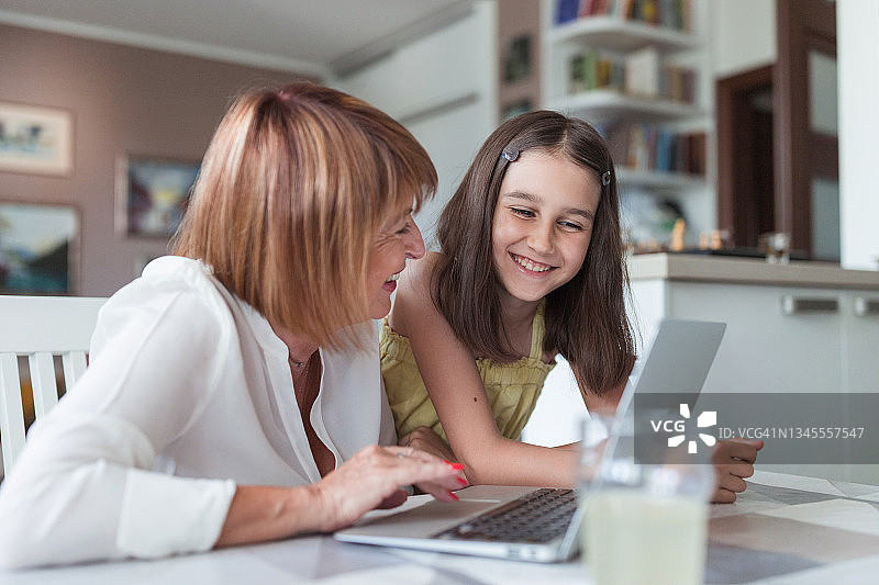 微笑的成熟女人使用笔记本电脑，观察坐在她旁边的快乐小女孩图片素材