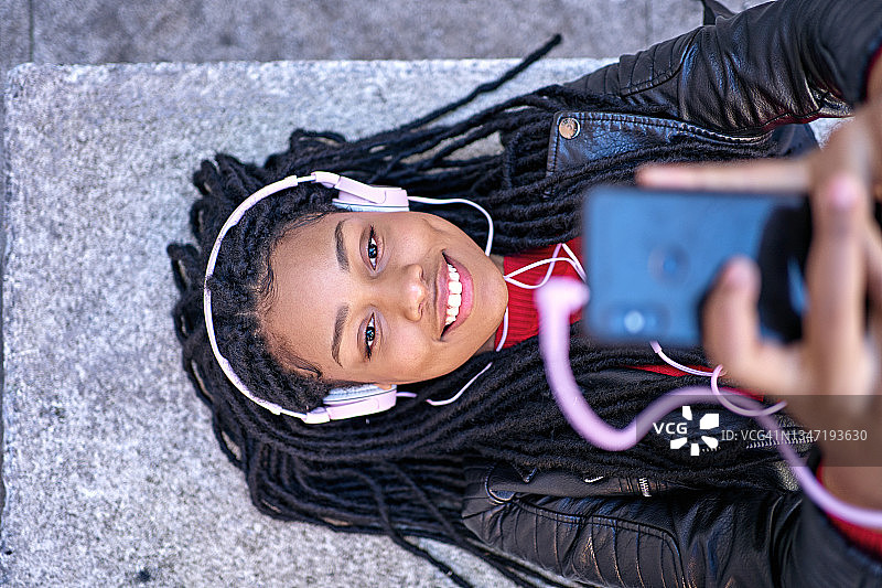 一名非裔美国妇女微笑着拿着手机，躺在城市的长椅上聊天或听音乐。头发被编成了脏辫。图片素材