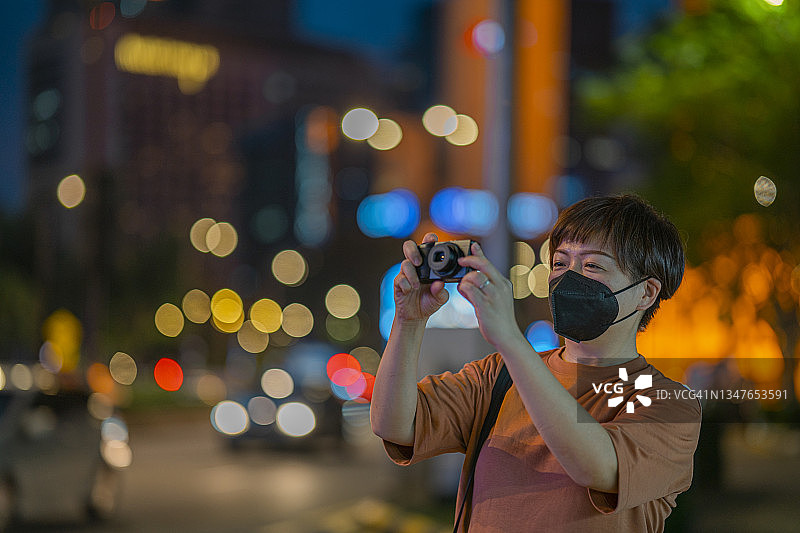 亚洲华人中年女游客带着面罩，在霓虹灯照亮的吉隆坡繁华街道上用相机拍照图片素材