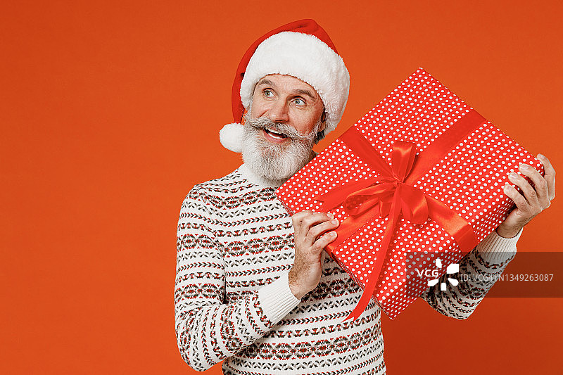 满脸大胡子的圣诞老人50多岁，戴着圣诞帽，穿着毛衣，摆着姿势，摆着展示，在素色背景的画室里孤立地放着纸箱。祝2022年新年快乐，庆祝圣诞佳节快乐。图片素材