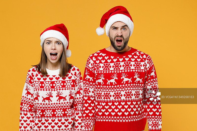 震惊而焦虑的年轻圣诞老人夫妇朋友男人女人穿着红色毛衣圣诞帽保持嘴巴张开孤立在黄色背景摄影棚肖像。新年庆祝快乐节日概念。图片素材