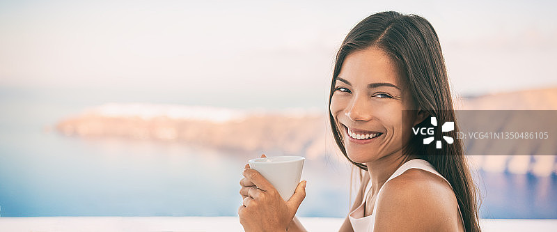 希腊伊亚岛，一名亚洲女子在豪华邮轮旅行度假室外餐厅享用早餐时喝咖啡，俯瞰地中海。全景横幅。图片素材