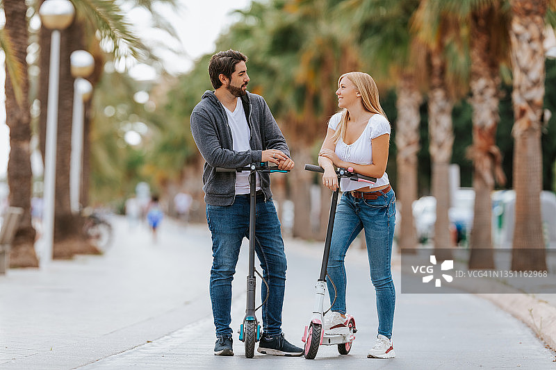 一对微笑的情侣在城市里用电动滑板车和生态交通工具互相看着图片素材
