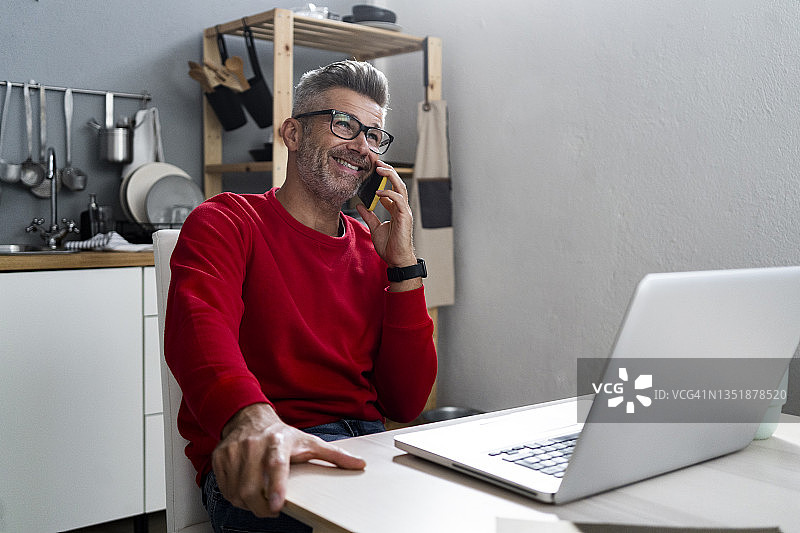 微笑的男人在家里用笔记本电脑和智能手机通话图片素材