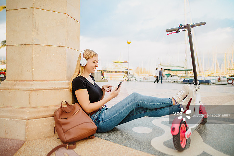 年轻的金发女子坐在港口，戴着蓝牙耳机，骑着电动车，用手机上网图片素材