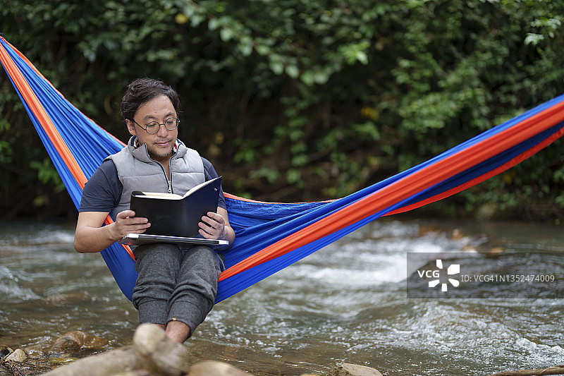 一名亚洲日本男子坐在森林露营地小溪边的吊床上看书。图片素材