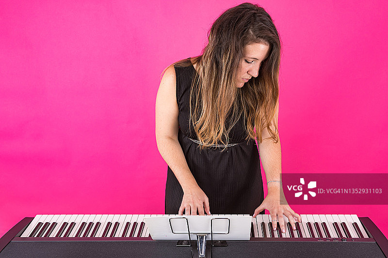 钢琴家音乐家女孩身穿黑色连衣裙，戴着带有高音谱号的戒指，在粉红色的背景上弹奏钢琴。音乐，钢琴家和作曲家的概念。图片素材