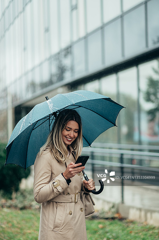 女商人打着雨伞，在下雨的时候用智能手机走在城市街道上图片素材