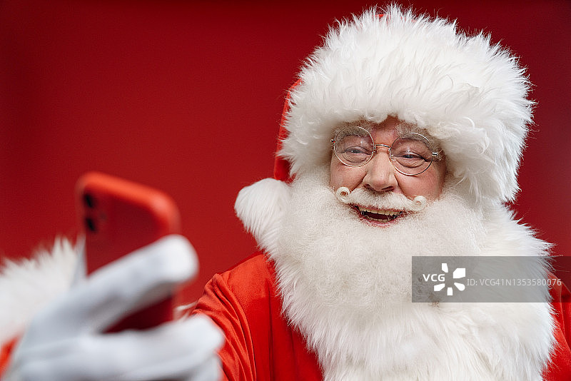 照片中的圣诞老人笑着看着他的红色智能手机，背景是明亮的红色图片素材